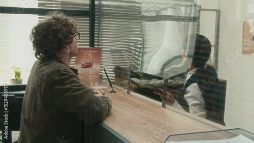 Female bank teller assisting male client with filling out documents checking customers passport at service window