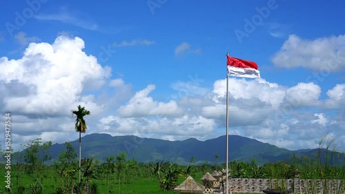 The red and white flag fluttering in the wind on a sunny day. Indonesian flag with blue sky, white clouds and mountains in the background.
