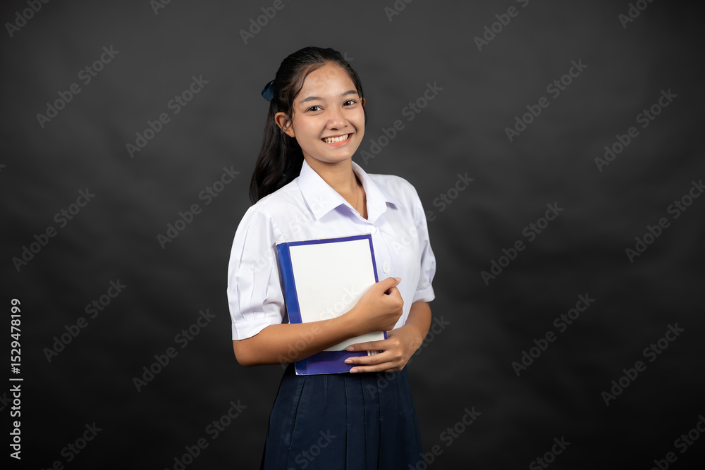 Obraz premium An Asian high school girl poses in a studio background, holding a notebook from her school, imagining her bright future at a leading university where she plans to study science.