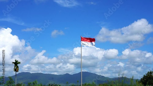 The red and white flag fluttering in the wind on a sunny day. Indonesian flag with blue sky, white clouds and mountains in the background.