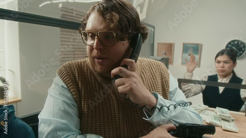 Medium shot of armed unprofessional male criminal discussing demands with female crime partner speaking to police on phone while counting wads of money at tellers counter during bank robbery