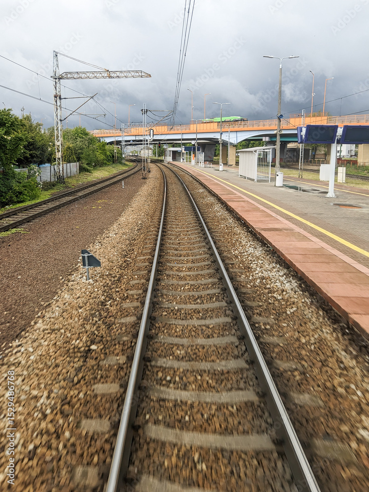 Naklejka premium Curved railway tracks leading out of a modern train station, captured from the rear of a moving train under dramatic stormy skies — symbolizing travel, transition, and the power of modern rail infrast