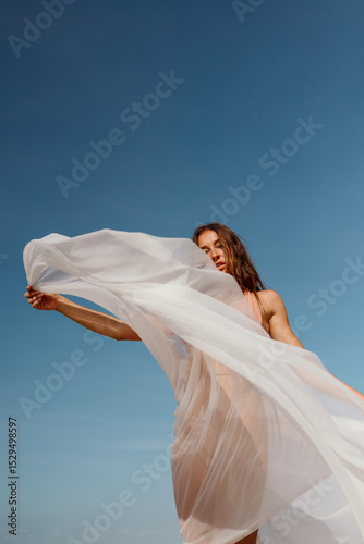 A sheer veil billows dramatically in the wind as a woman holds it high against a bright blue sky. A minimalist visual of grace and movement.