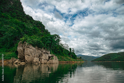 Fototapet Panoramic view of a tranquil lake reflecting an overcast sky, with dense tropical forest clinging to steep hillsides and a prominent rock formation
