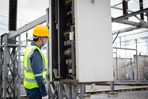 Engineer working at high voltage substation inspecting electrical control panel with safety helmet and reflective vest in industrial outdoor environment