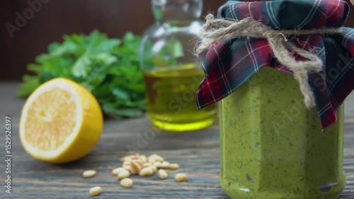 A hand puts a glass jar with fresh green pesto on a wooden table. In the background are ingredients for this healthy homemade Italian sauce like basil pine nuts garlic and cheese