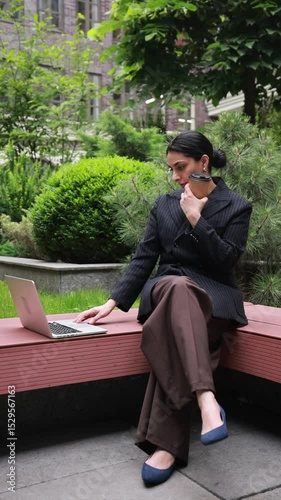Confident young woman in black business suit sits on bench in office yard, typing on laptop, focused on work. Concept of workplace flexibility, career development, remote working.