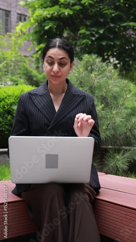 Professional woman in formal black attire seated near corporate building, speaking on video call via laptop with serious expression. Concept of business conversation, online meeting, trust.