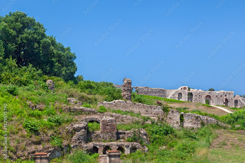 Fototapeta premium Ancient ruins of Nikopolis in Epirus, Greece, under a clear summer sky. Historic Roman city overgrown with vegetation and scattered remains.
