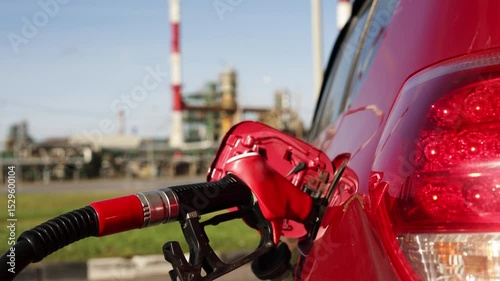 Gas pump nozzle inserted into red car fuel tank at oil refinery background, symbolizing gasoline refueling and energy industry. Gas pump nozzle filling gasoline car