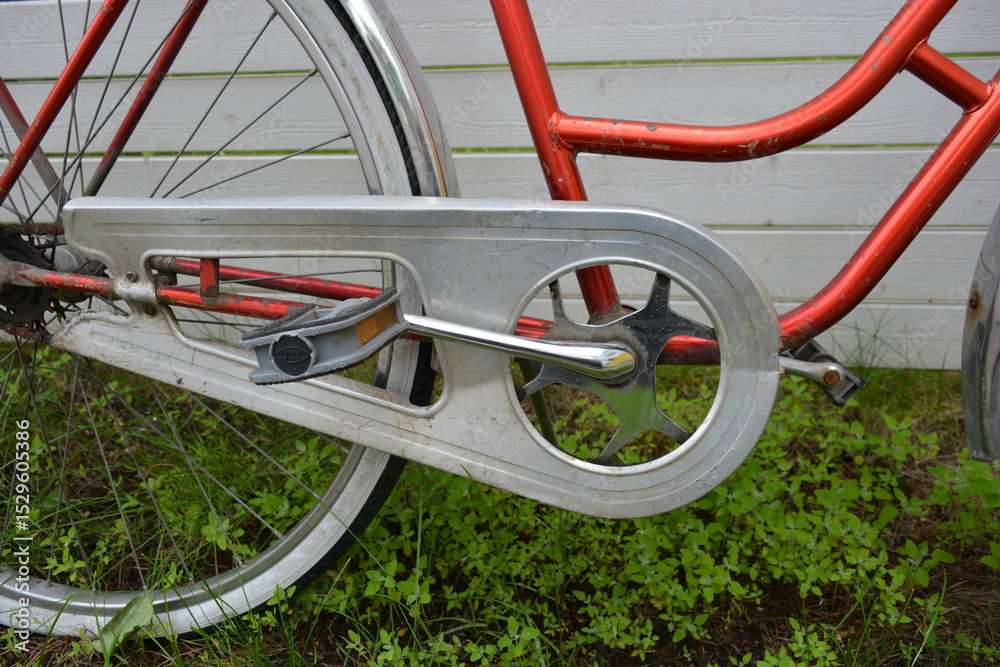 An old red metal bicycle stands against a white fence, on a green lawn. The bicycle parts are old, the bicycle wheels are rusty, there are many red spots of rust on the metal parts of the bicycle. An old red metal bicycle stands against a white fence, on a green lawn. The bicycle parts are old, the bicycle wheels are rusty, there are many red spots of rust on the metal parts of the bicycle.