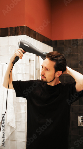 Young man drying hair with a hairdryer in bathroom