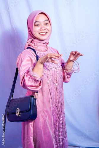 Asian Muslim woman wearing pink baju kurung with sparkling embroidery details smiling at camera with sling bag on her shoulder.
