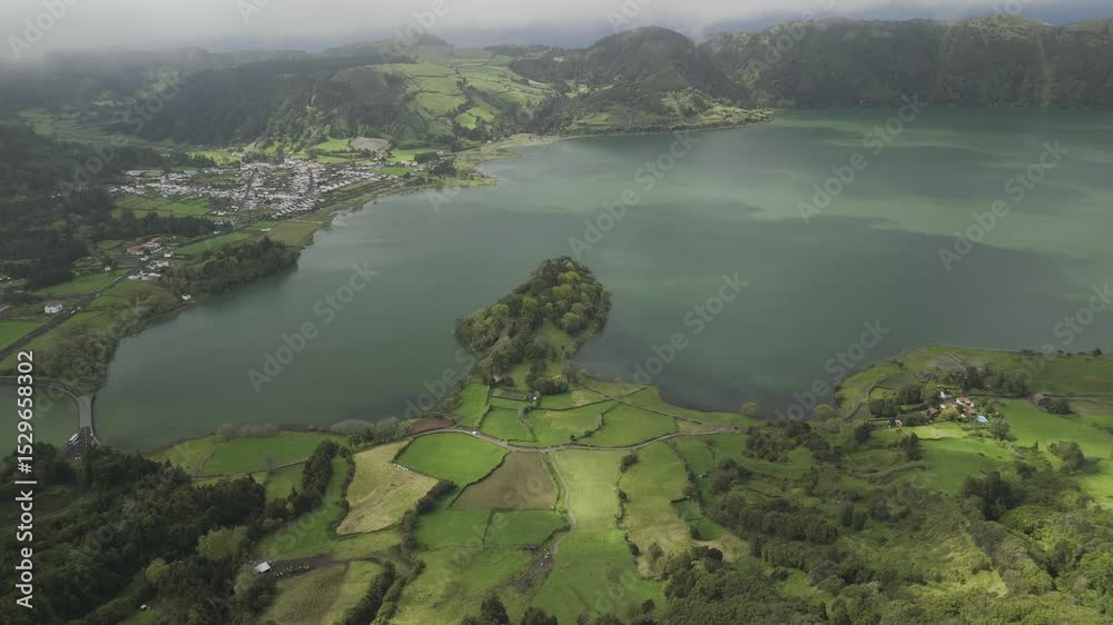 Drone flies toward peninsula jutting into lake with town across the water in Sete Cidades, Sao Miguel, Azores, Portugal