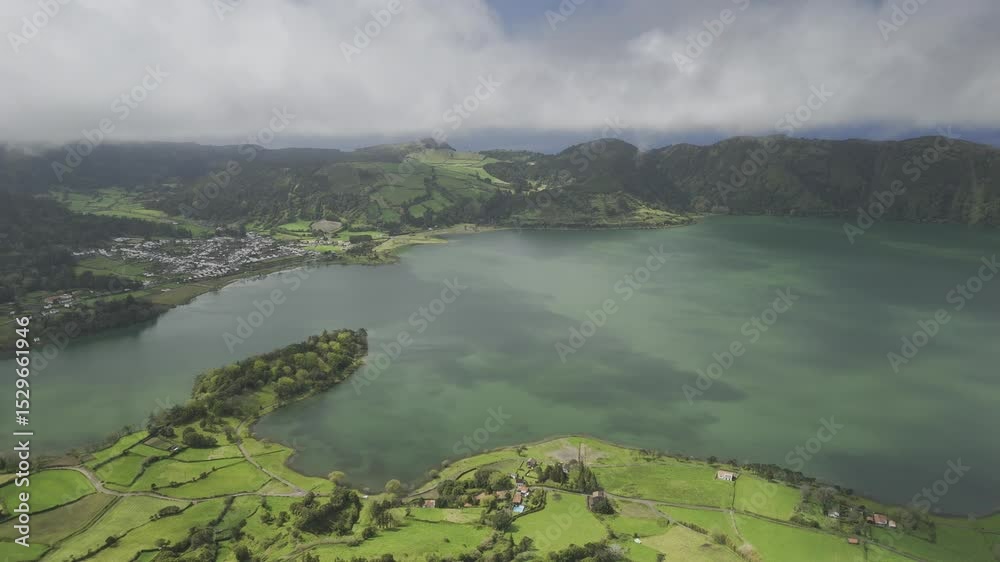 Drone flies to the left high over Lagoa Azul with a small town inside the volcano crater in Sete Cidades, Sao Miguel, Azores, Portugal