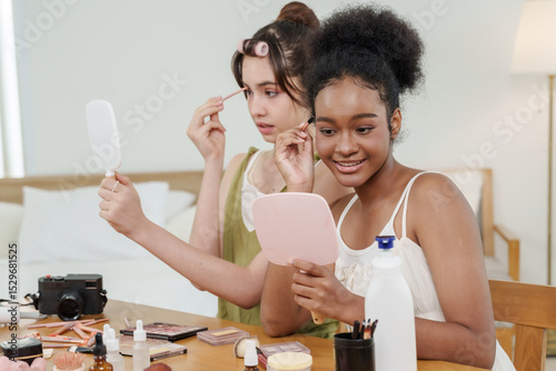 african female teen and asian female teen smile while applying eyeliner in front of mirror during makeup tutorial for content creation in warm bedroom setup with cosmetics