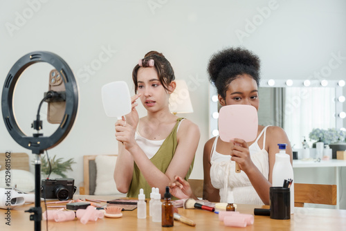 asian female teen and african female teen sit at table applying makeup while looking into mirrors and recording online beauty tutorial together with cosmetics and tools spread across surface