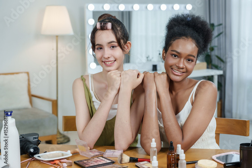 asian female teen and african female teen pose with hands together smiling after skincare session during online content creation in bright beauty studio with cosmetic items on table