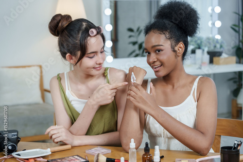asian female teen and african female teen show applied cream on arm to smartphone camera while smiling during beauty product review filming with cosmetics and tools on desk