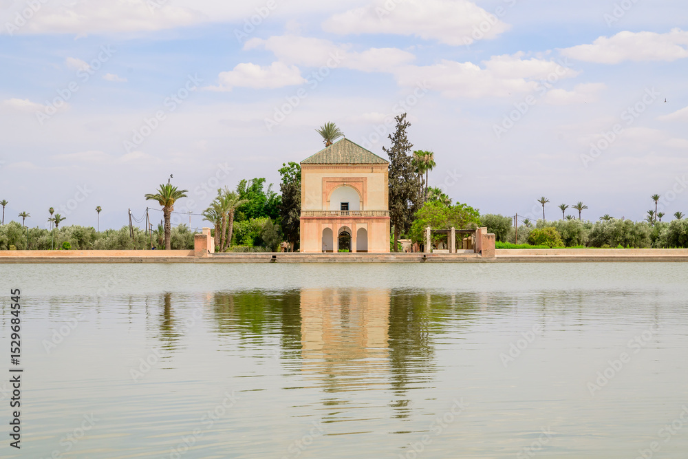 Obraz premium The Menara gardens Historic pavilion in Marrakech with reflections in a serene lake. Palm trees and blue sky enhance the tranquil setting. Ideal for travel and architecture themes.
