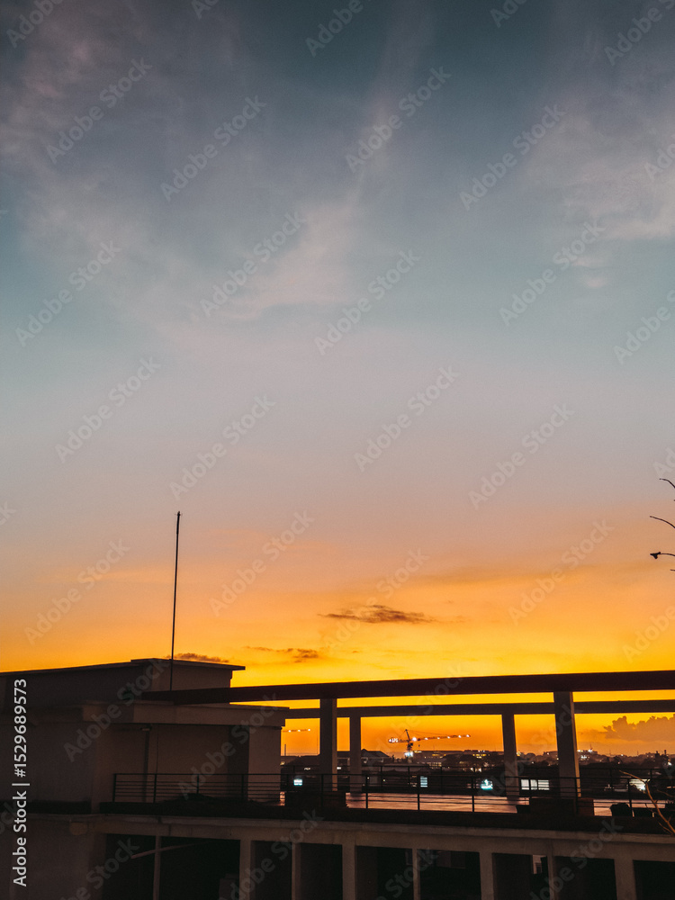 Obraz premium A vibrant sunset bathes the sky in hues of orange and pink, casting a warm glow over silhouetted buildings, creating a serene and picturesque urban skyline.