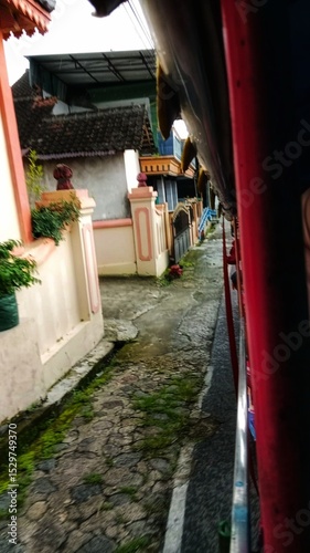 Fototapeta Naklejka Na Ścianę i Meble -  A narrow street in a quiet residential area lined with houses and small buildings, creating a cozy neighborhood atmosphere under a clear sky.
