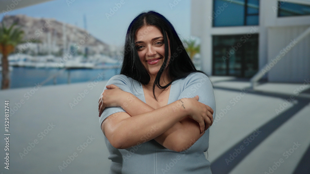 Fototapeta premium Woman embracing herself with a joyful expression, standing on a sunny seaside promenade with boats and hills in the background, capturing a serene and uplifting outdoor moment.