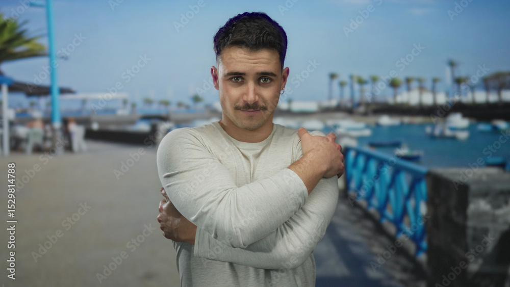 Fototapeta premium Young hispanic man embracing himself on a seaside promenade, with a serene ocean backdrop and clear blue skies, providing a peaceful outdoor setting.