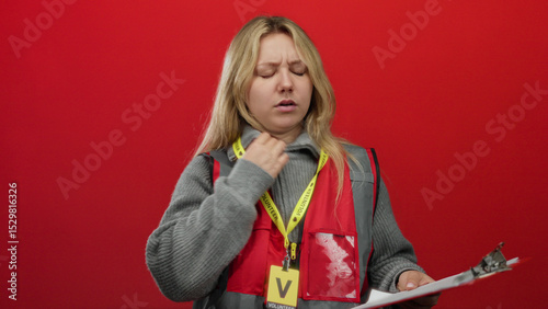 Young volunteer woman with blonde hair stands against an isolated red background wearing a reflective vest and holding a clipboard.