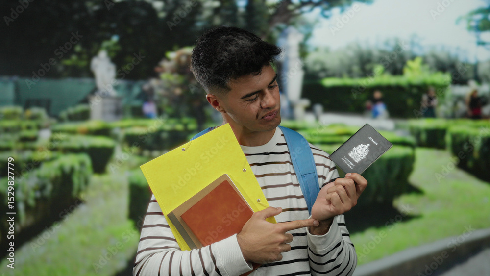 Fototapeta premium Man holds canadian passport and documents in a park, wearing striped shirt and blue backpack while pointing at the passport with a thoughtful expression.