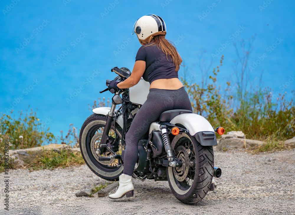 Obraz premium A rear view of a young woman on her customized chopper-style motorcycle parked at a coastal viewing point, looking at her mobile phone attached to the handlebars with the sea in the background.