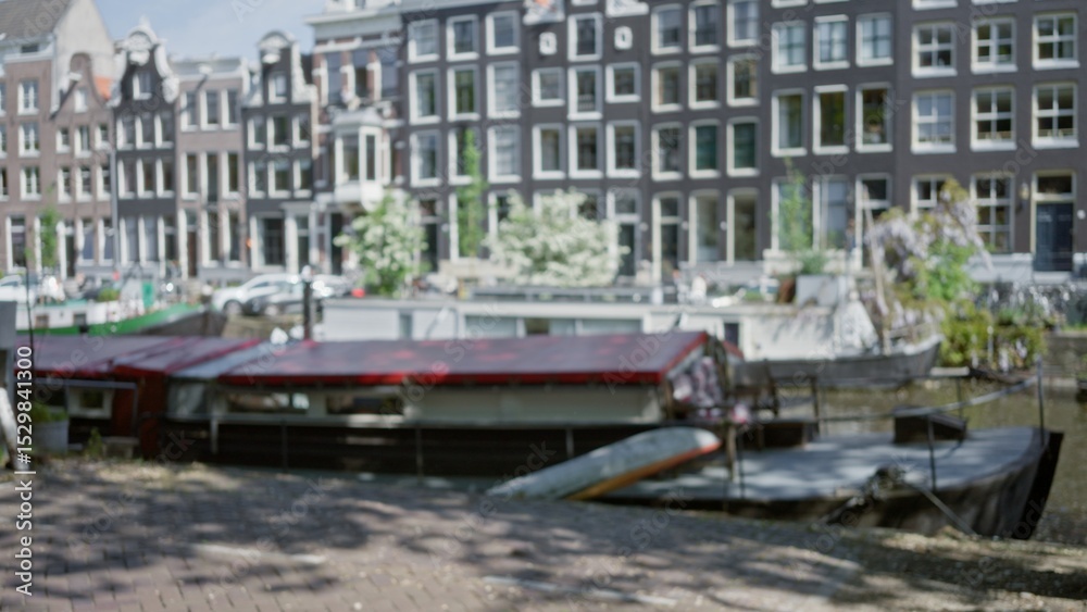 Naklejka premium Blurred image of amsterdam canal with houseboats moored under bokeh effect, featuring iconic dutch architecture in background under sunny sky, creating a serene urban scene.