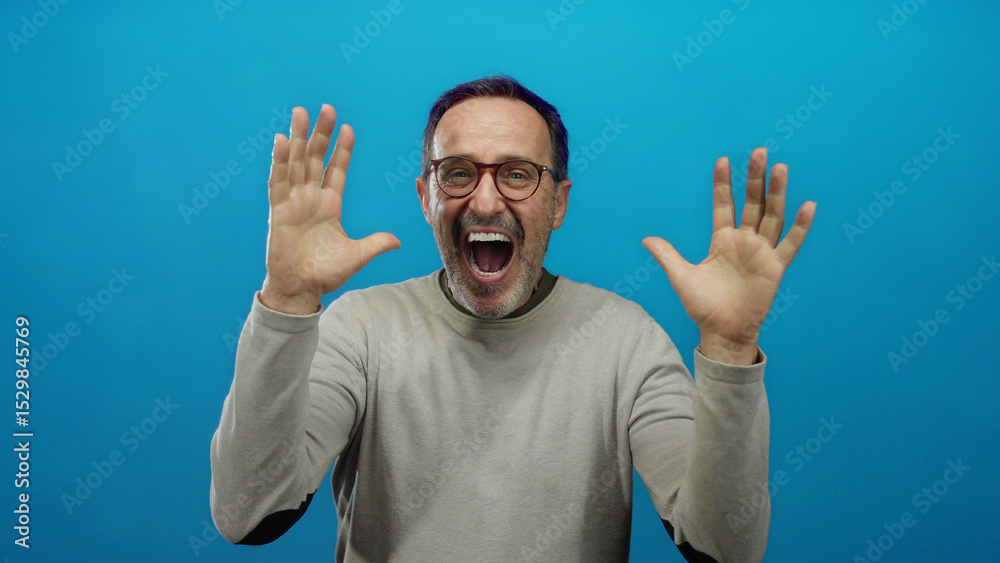 Naklejka premium Mature hispanic man expressing joy and excitement with raised hands against a vibrant blue background, showcasing an enthusiastic and animated expression.