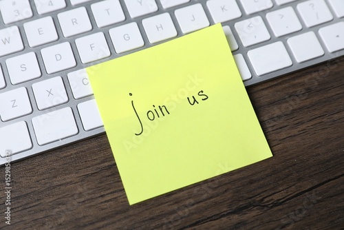 Fotografie Paper note with phrase Join Us and keyboard on wooden table, closeup