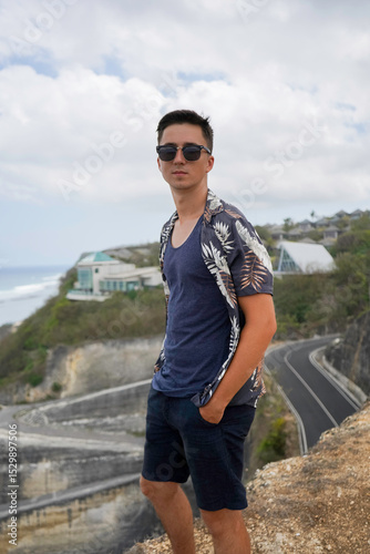 Wallpaper Mural A young man in sunglasses and a tropical shirt stands on a cliff overlooking the ocean and Melasti beach on a cloudy day in Bali, Indonesia Torontodigital.ca