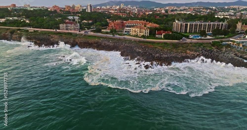 Aerial view hugging rocky coast by the sea in Cascais, Portugal with waves crashing underneath