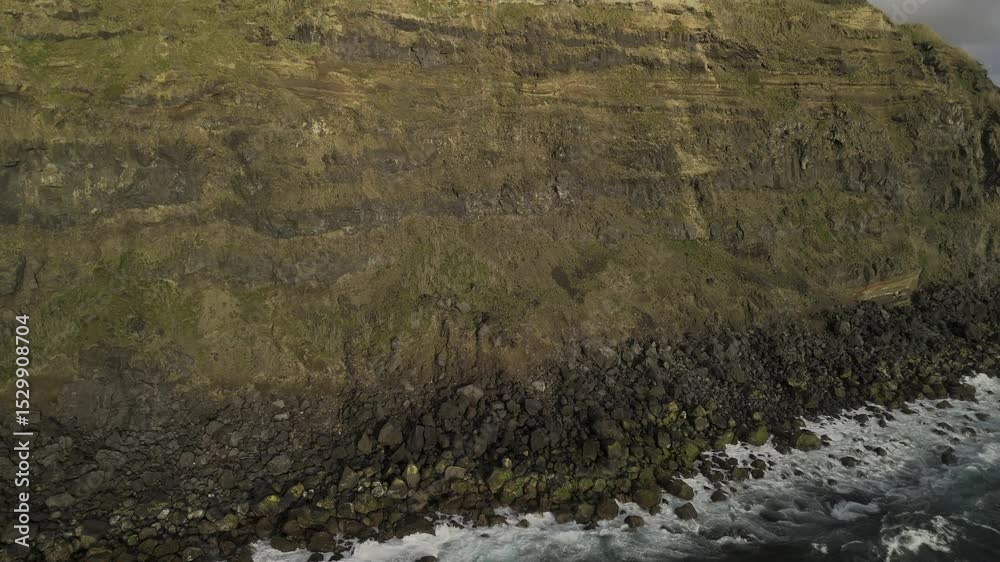 Drone rises up from sea in front of sunset-kissed cliffs of Ponta da Ferraria to find a lighthouse in the middle of expansive green fields in Sete Cidades region, Sao Miguel, Azores, Portugal