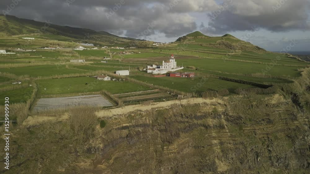 Drone flies inland from sunset-kissed cliffs over red and white lighthouse on Ponta da Ferraria in Sete Cidades region, Sao Miguel, Azores, Portugal