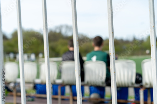 View of players on the bench observing a soccer match through a fence at a sports field during daylight hours