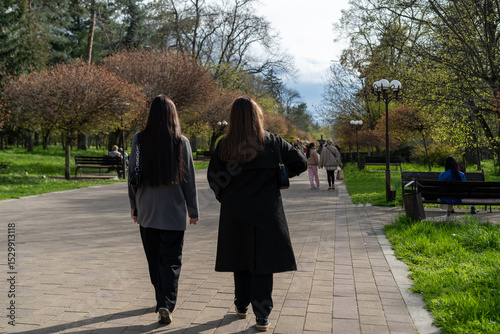 Two people stroll through a public park lined with trees and benches on a sunny day in springtime