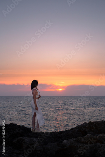 A young girl in a white dress poses backlit at sunset at Cape Artrutx on the Balearic island of Menorca.