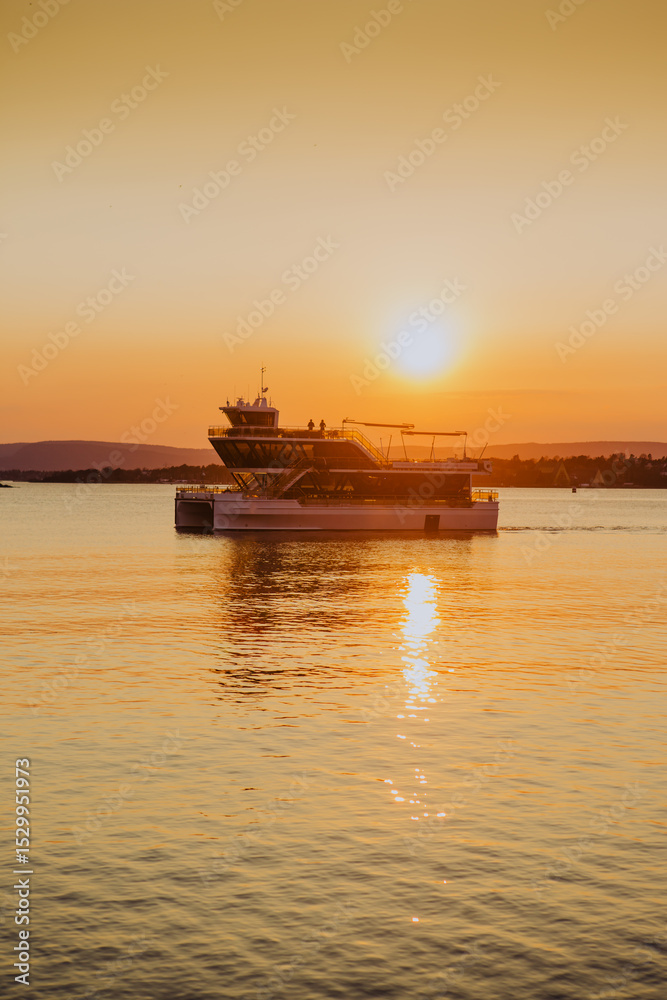 Obraz premium Bathed in the golden light of sunset, a local ferry glides across Oslo harbour, creating a warm and inviting scene against the city's waterfront