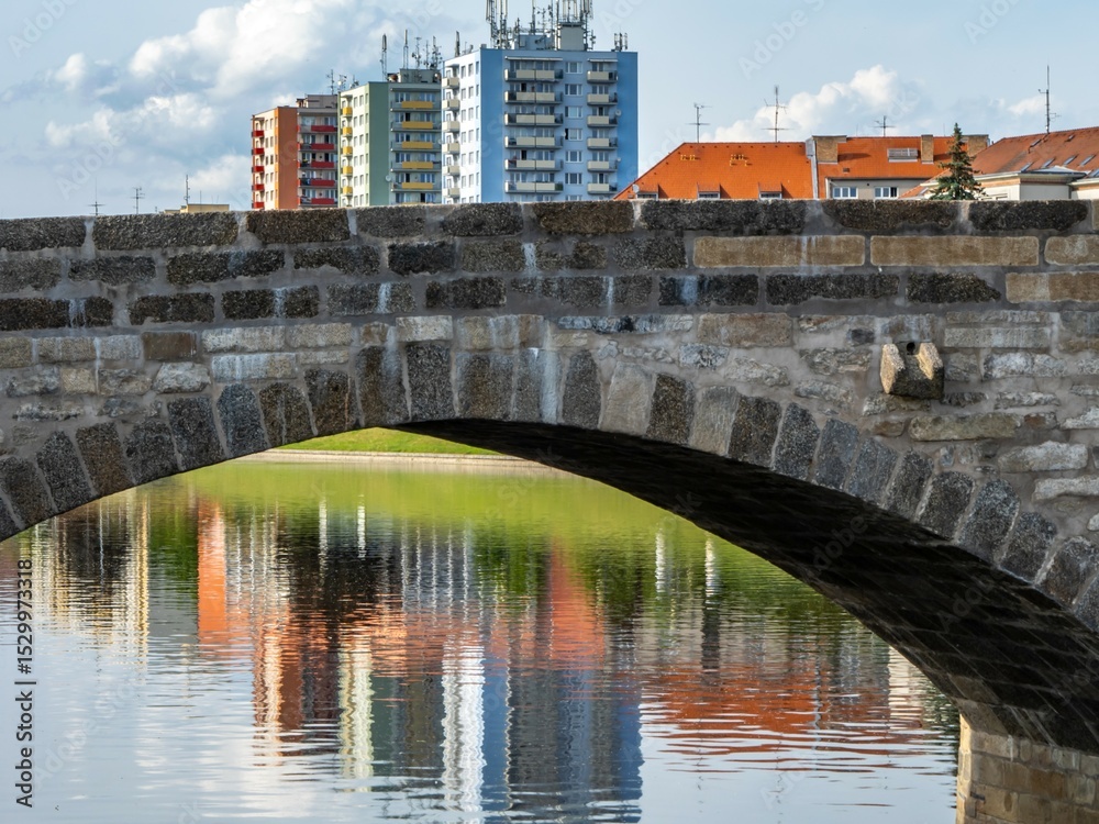 Naklejka premium Colorful apartment buildings reflected in calm water beneath a stone bridge arch. Urban landscape with contrast between historical architecture and modern housing