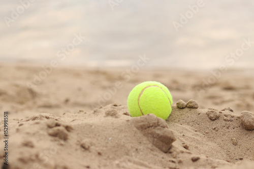 Yellow tennis ball lies on the sand. Sports and recreation concept, summer games. Tennis or paddleball ball. Tennis court near the beach. Paddleball ball on the sand near the water. Outdoor games