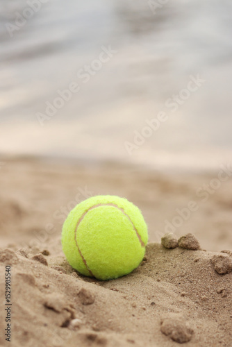 Yellow tennis ball lies on the sand. Sports and recreation concept, summer games. Tennis or paddleball ball. Tennis court near the beach. Paddleball ball on the sand near the water. Outdoor games