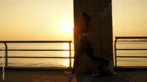 Jogger running along a waterfront path at sunrise, silhouette against orange sky