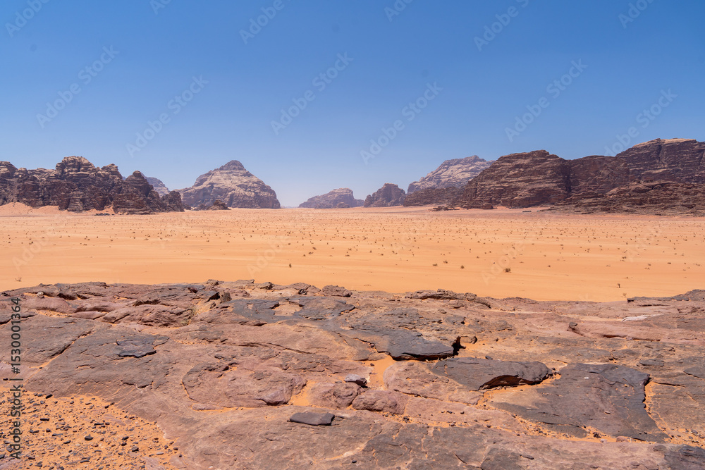 Naklejka premium Wadi Rum Desert Landscape: A panoramic view of the Wadi Rum desert in Jordan, showcasing vast expanse of red sand, rugged mountains, and clear blue sky.