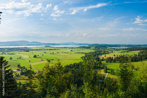 Scenic aerial view of wide open green fields, small farms, forests, and shimmering lakes under a blue sky with scattered clouds on a sunny day.