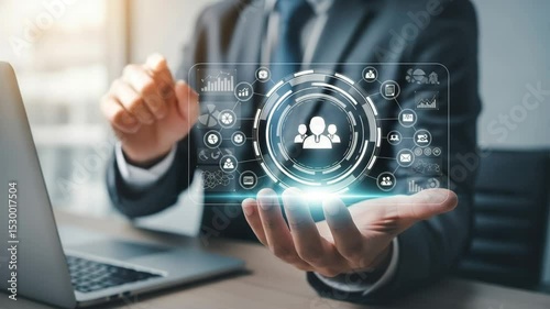 Man in suit holding a digital interface with team icon and laptop in blurred background view close up
