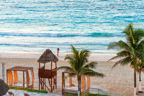Fototapeta Naklejka Na Ścianę i Meble -  Woman walking along a serene beach in Cancun, surrounded by palm trees, wooden cabanas, and gentle ocean waves High-quality photo